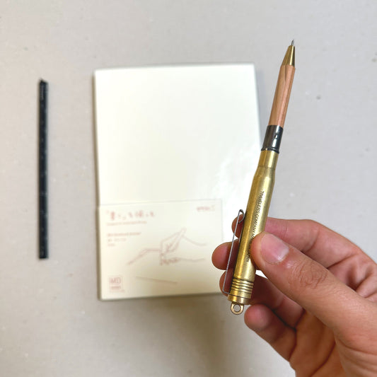 Mano che tiene una penna in ottone con patina, accanto a taccuini e strumenti da scrittura.